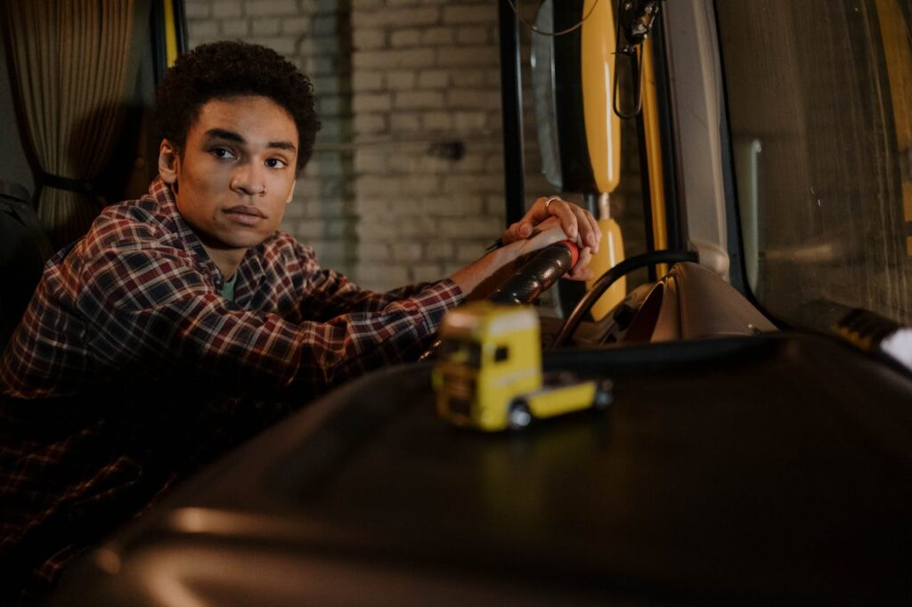 pexels-photo-7541982 A young man sits in the cabin of a truck, hands on the steering wheel, with a model truck on the dashboard.